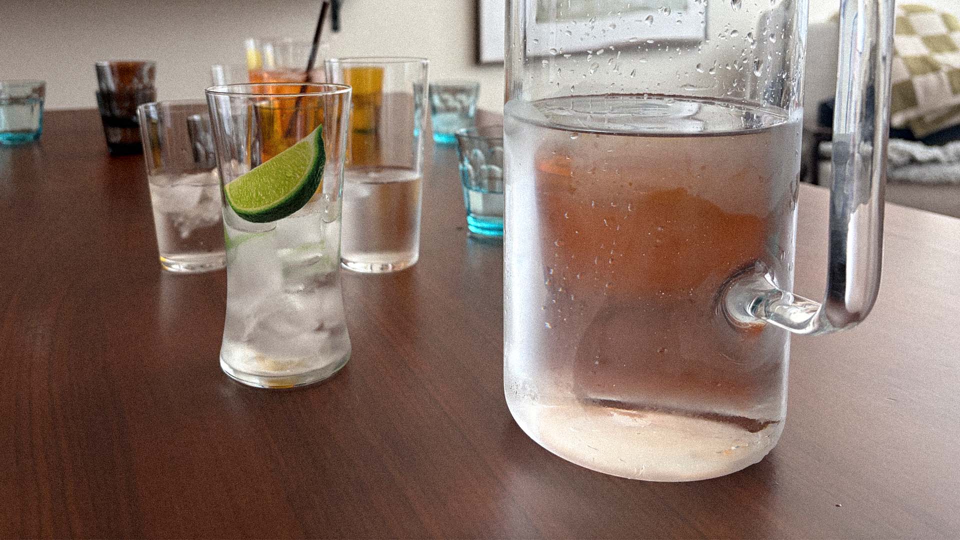 Glass pitcher and everyday drinking glasses gathered around a wooden table with ice water and a fresh lime wedge, capturing a quiet shared moment.
