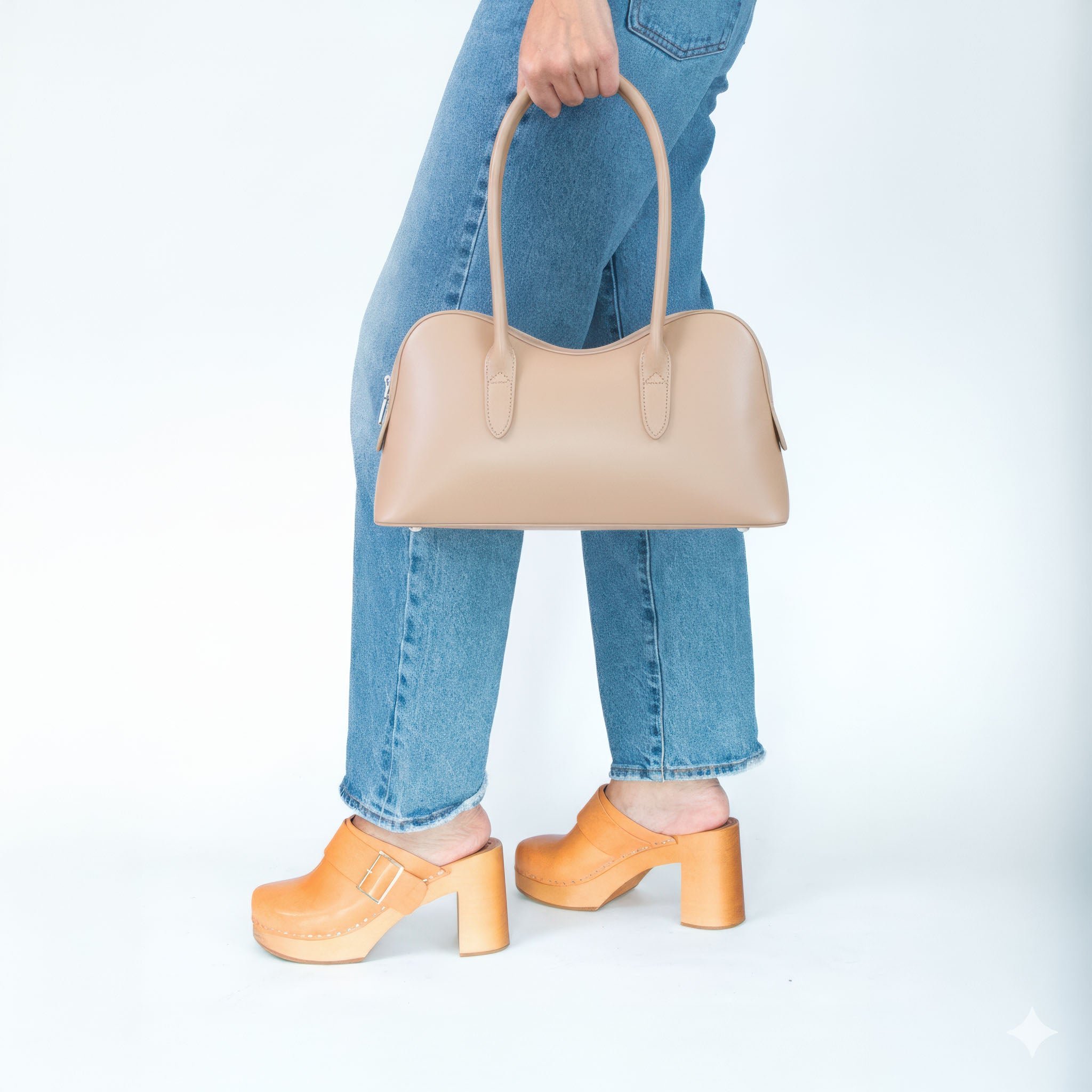 Person holding a nude handbag with blue jeans and saddle colored shoes on a white background