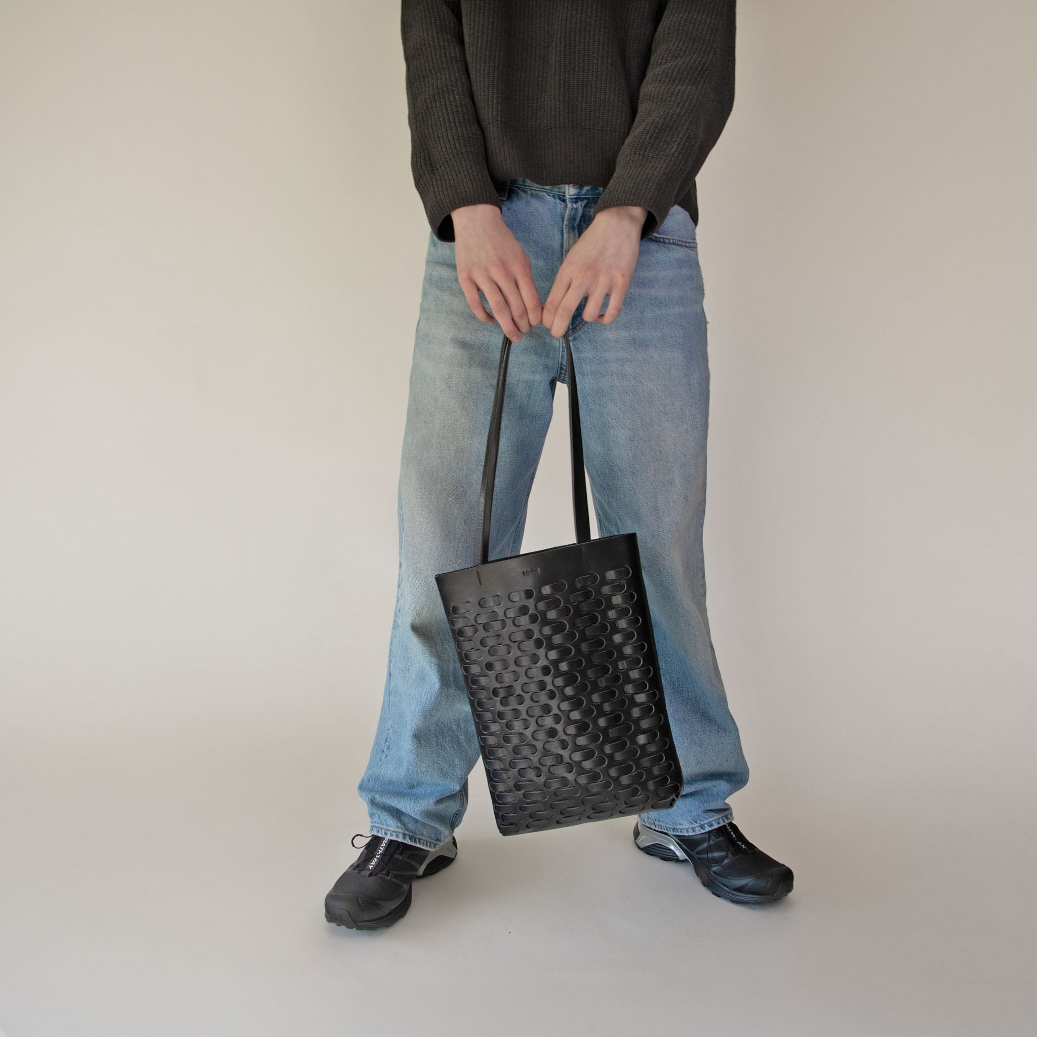 Person holding NImes black woven tote wearing blue jeans and black shoes on a plain background