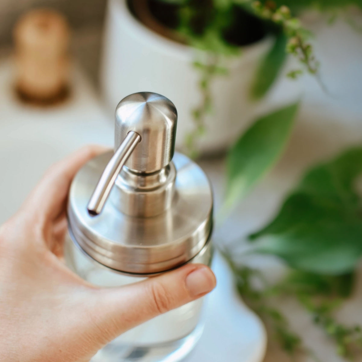 A hand holding a clear glass soap dispenser with a brushed stainless steel pump and lever, set against a softly blurred bathroom or kitchen background with green plants.