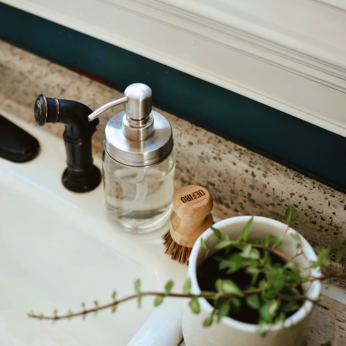 A clear glass soap dispenser with a brushed silver pump and curved spout, styled on a speckled stone countertop next to a black faucet, a small wooden scrubber, and a potted green plant.