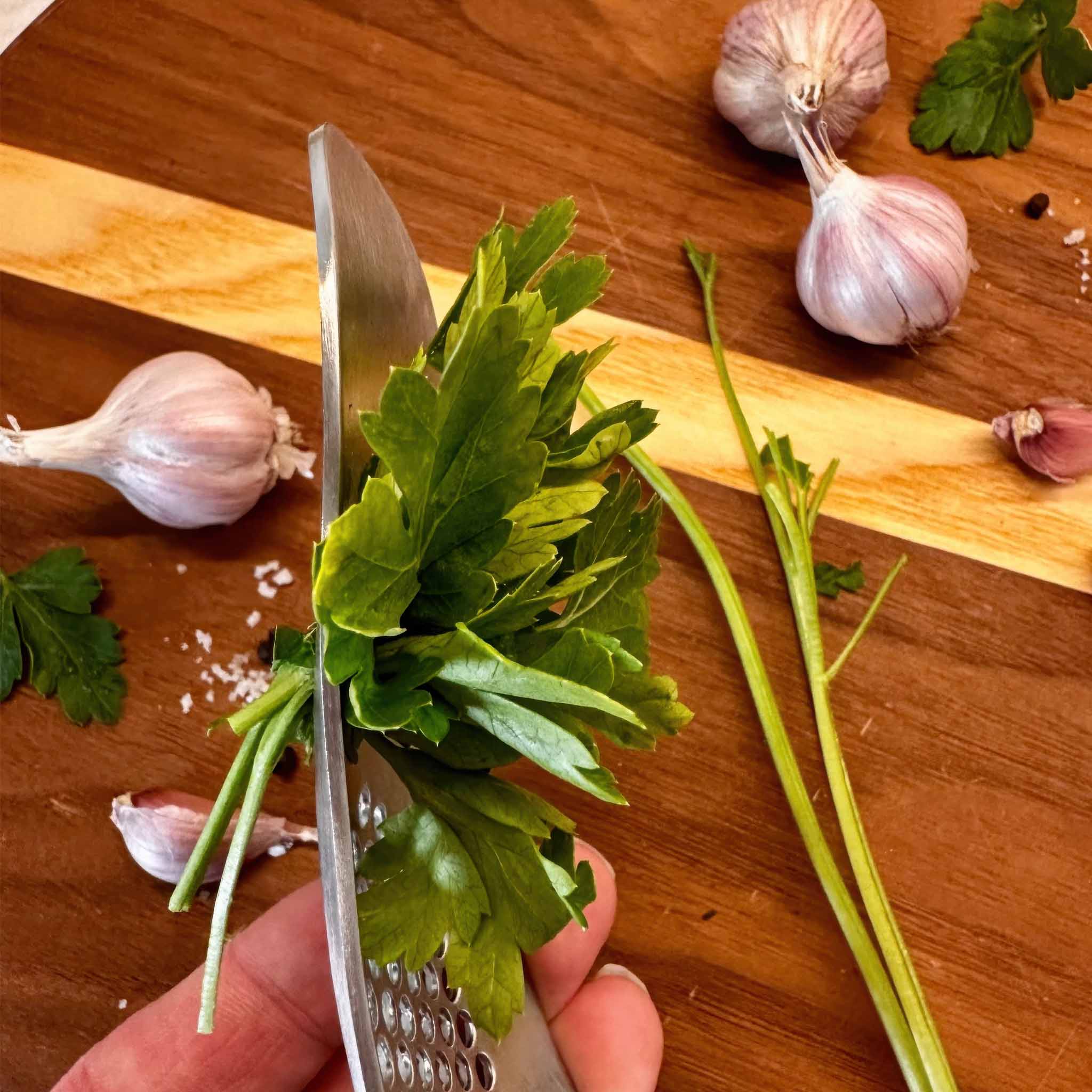Person stripping green herbs with the herb/garlic press on a wooden cutting board with garlic cloves.