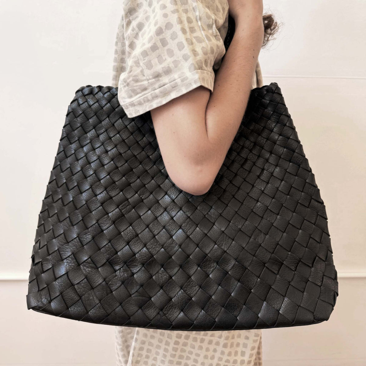 A person wearing a beige patterned top carries a large black woven leather tote bag against a plain light background.