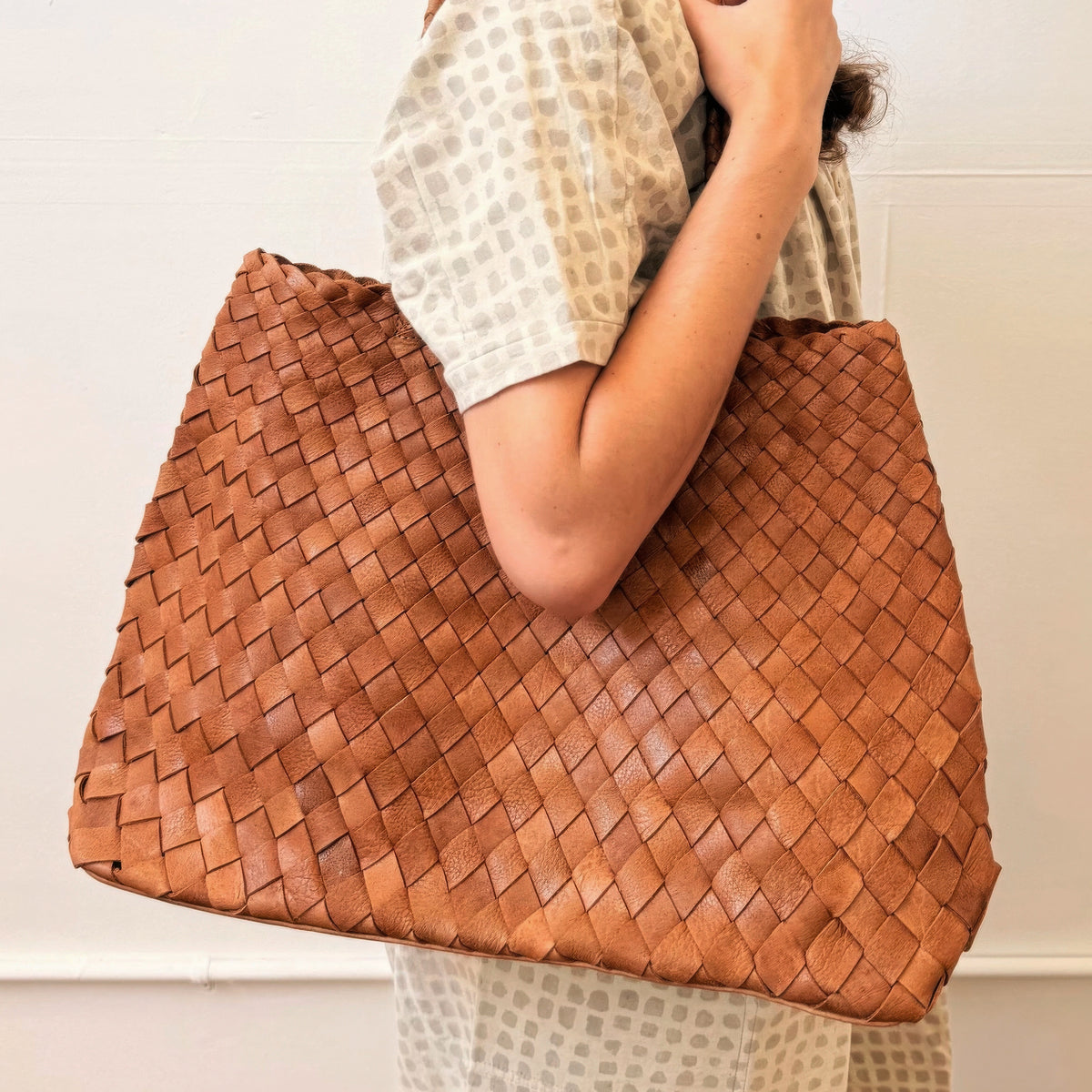 A person wearing a beige patterned top carries a large woven leather tote bag in a warm tan-brown color against a plain off-white wall.