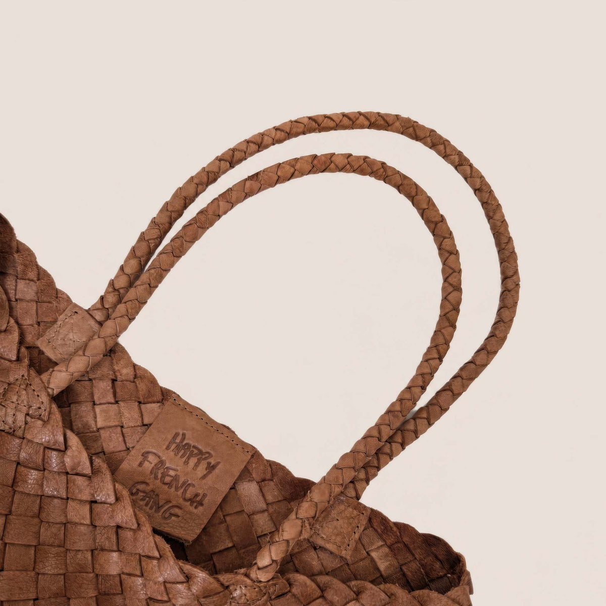 Close-up of a woven brown leather tote bag with braided shoulder straps and a small leather tag that reads 'HAPPY FRENCH GANG' on a light beige background.