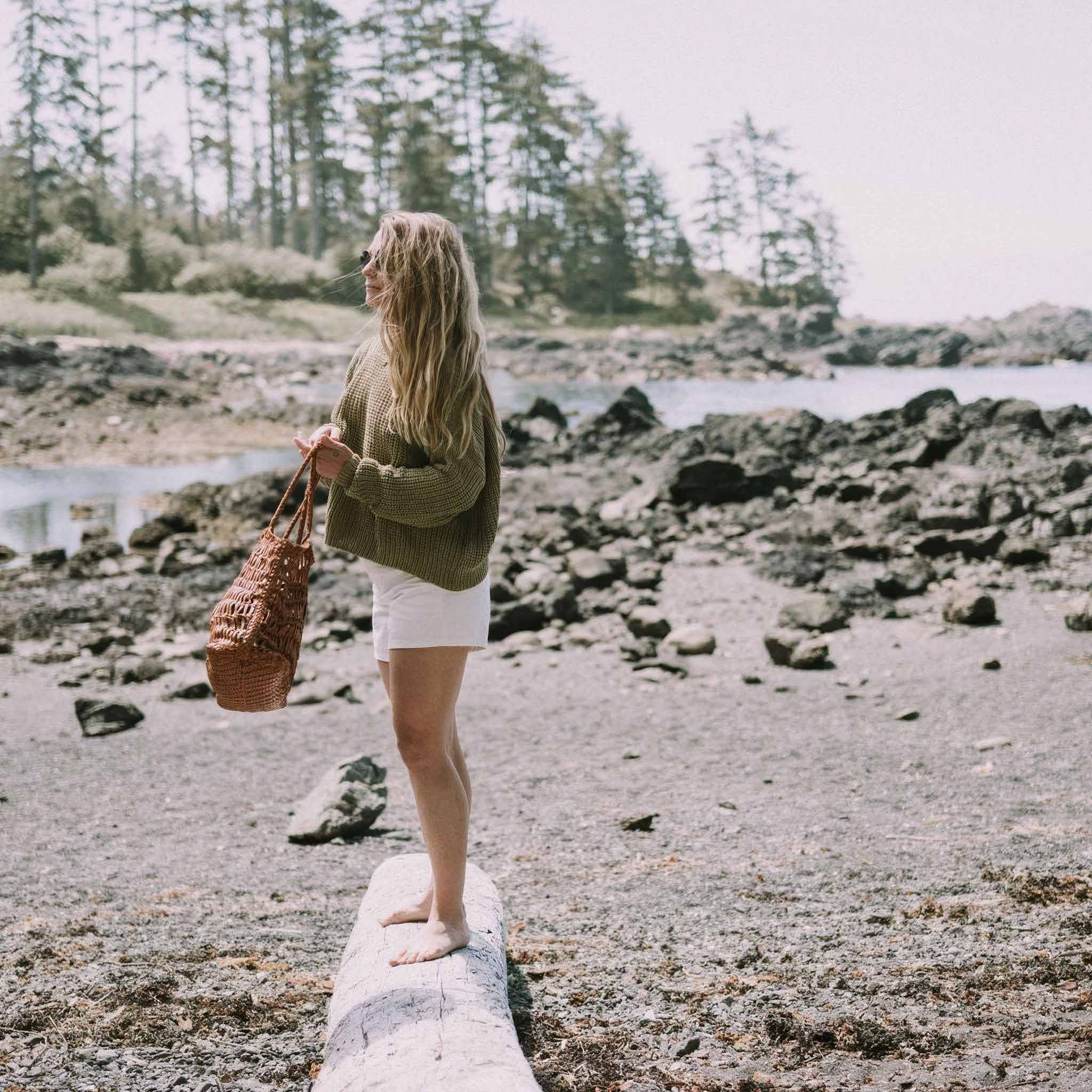 Woman carrying a leather weave bag on a rocky shoreline with trees in the background