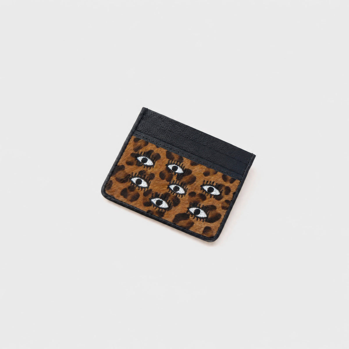A small card holder with a black top section and a brown leopard print lower section featuring embroidered eyes, shown on a light gray background.