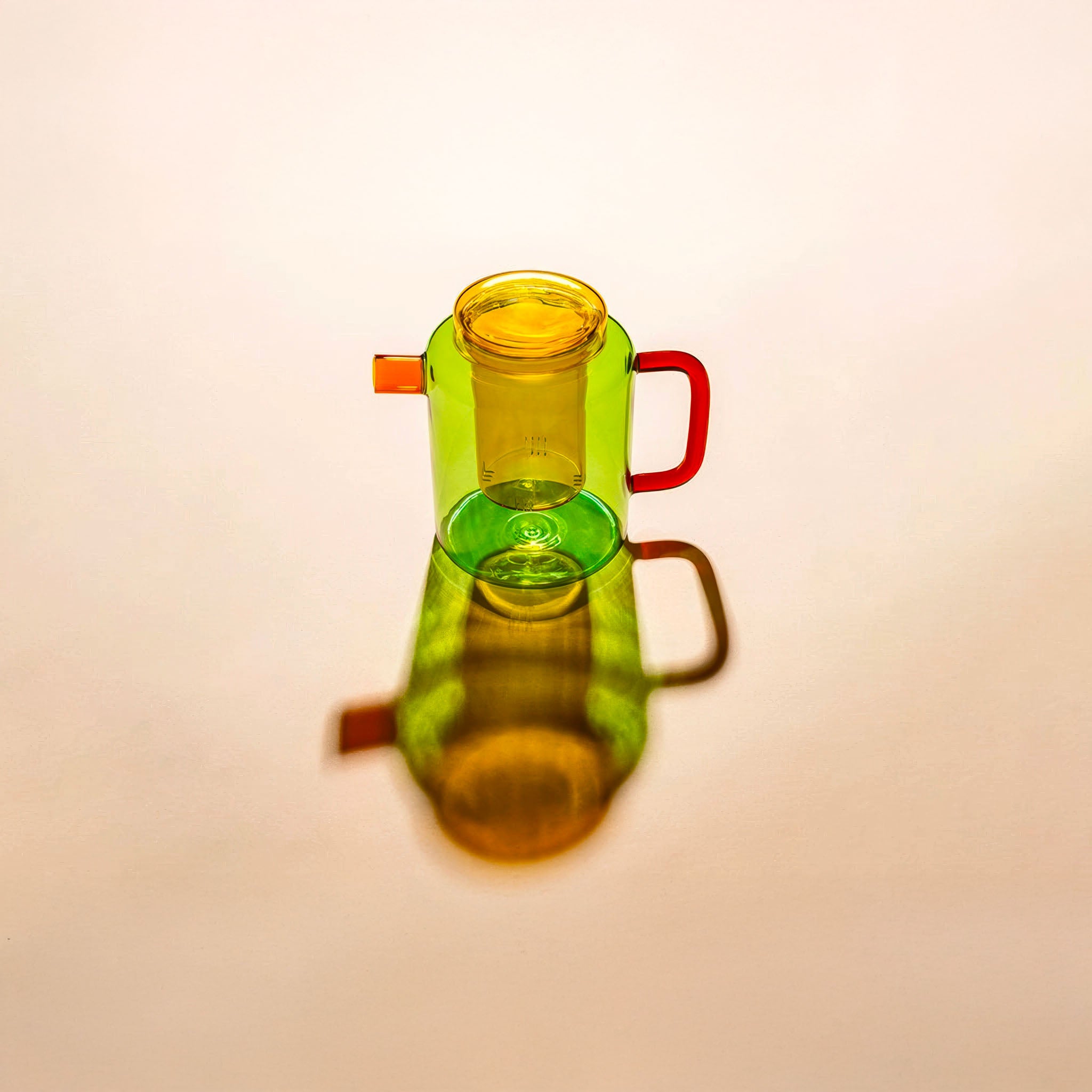 Colorful glass teapot with amber lid, yellow infuser, light green body, and red handle, shown against an amber background