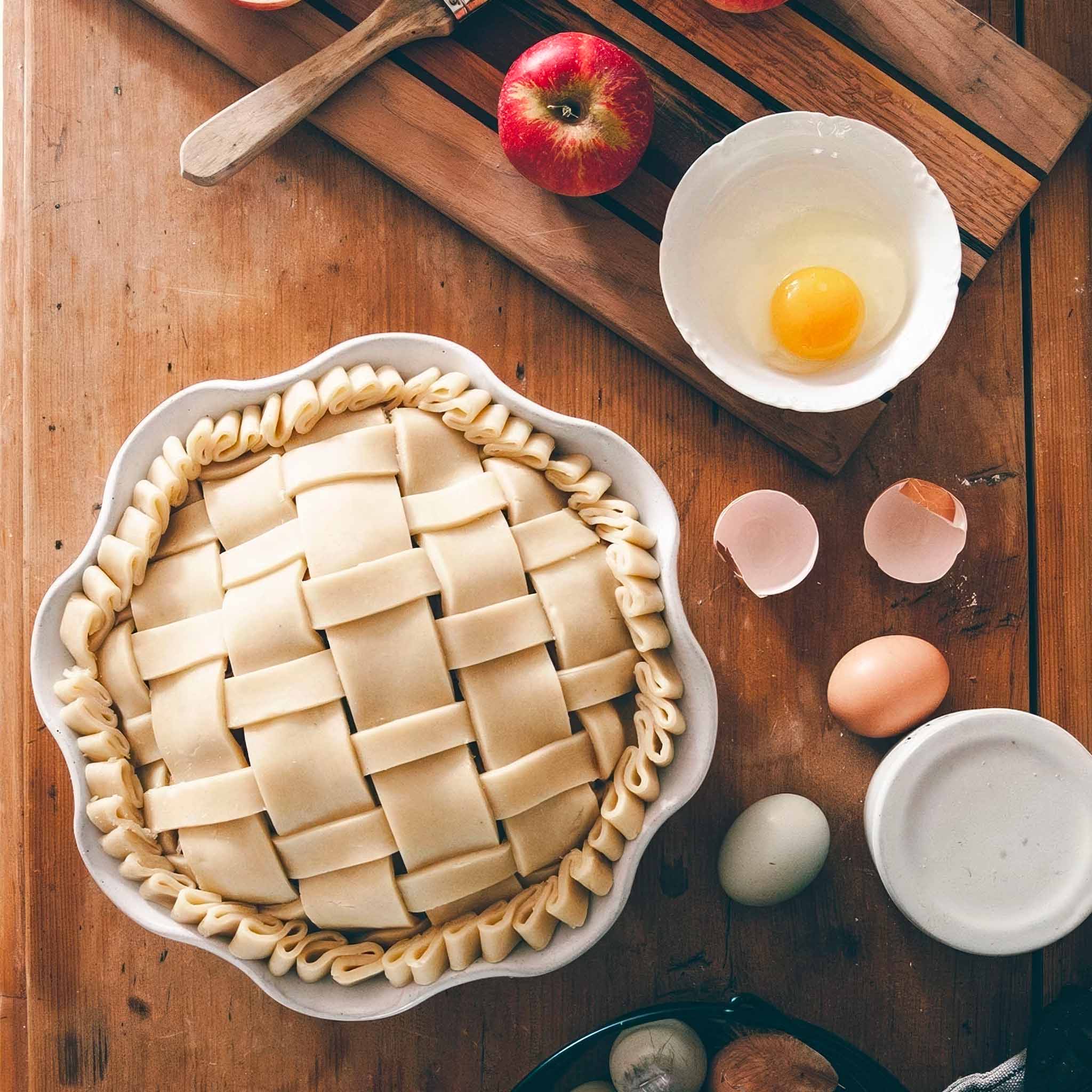 Unbaked lattice-topped pie i na white ceramic pie dish on a wooden table with eggs and an apple.