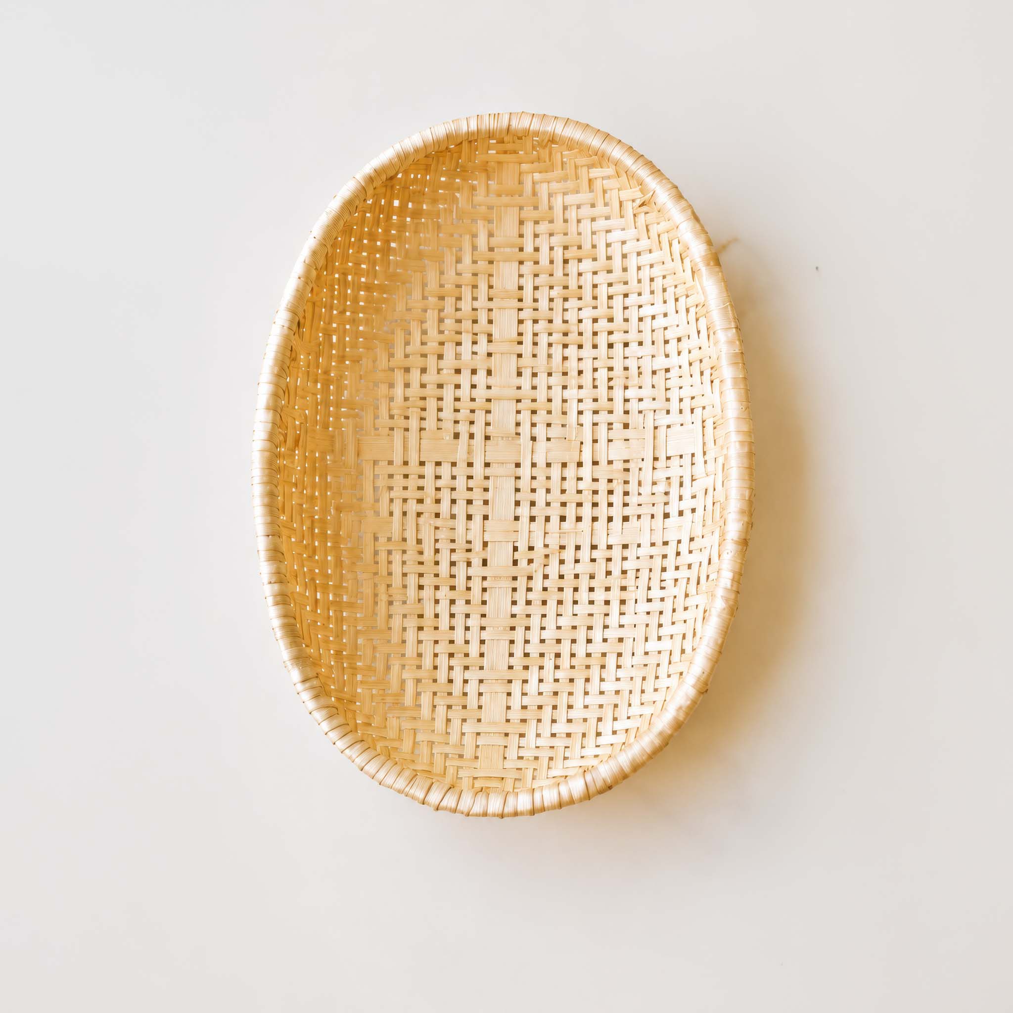 An oval woven rattan tray with a natural light tan color, shown from a top-down perspective on a clean, off-white background.