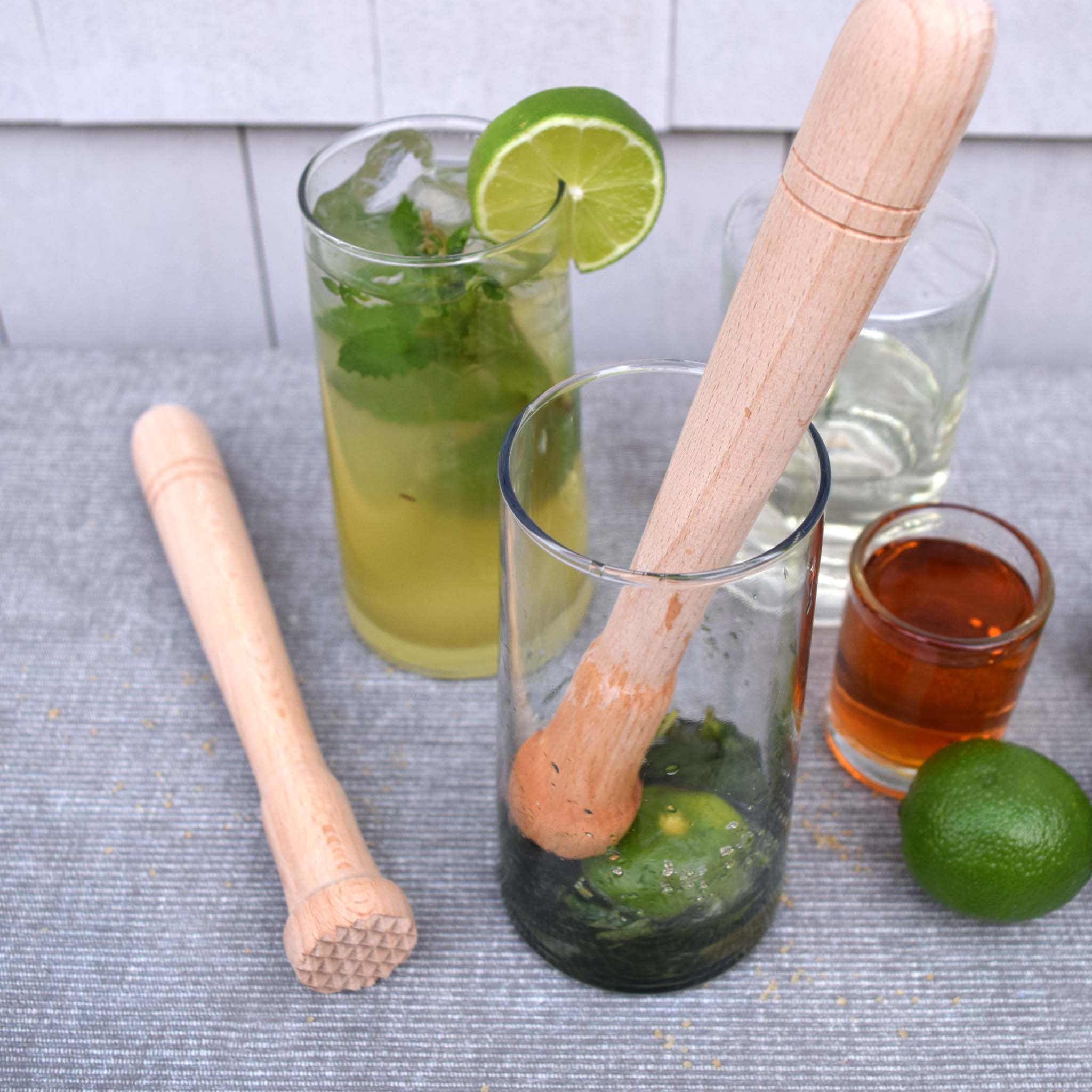 Muddler, glass with lime and mint, and other ingredients on a gray surface.