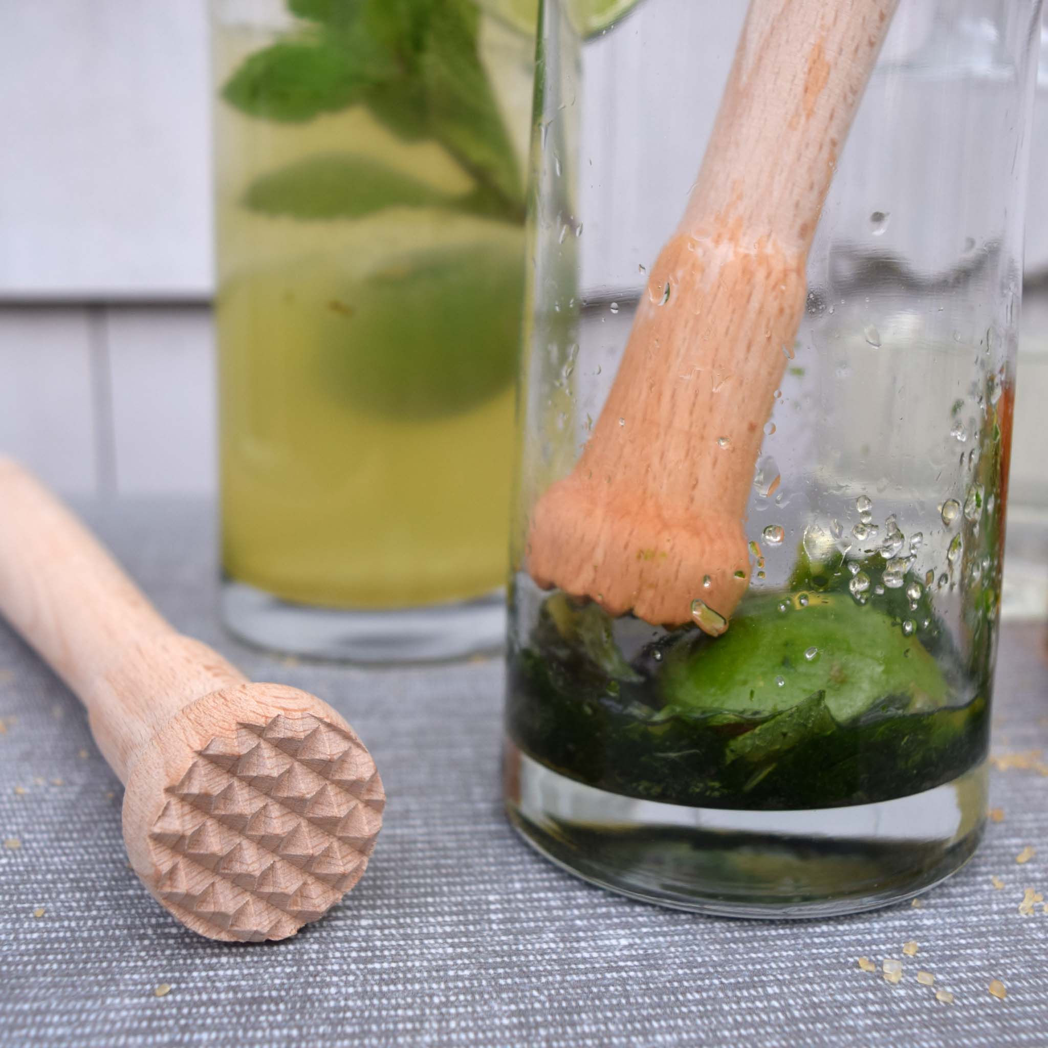 Glass with lime and mint leaves, wooden muddler, and another glass in the background on a gray surface.