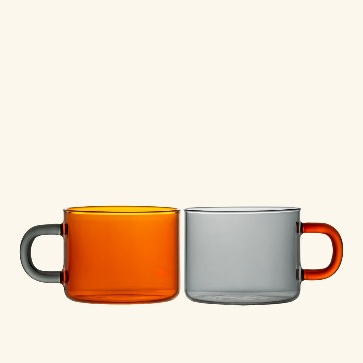 Two translucent glass mugs with thick handles on a light cream background. The mug on the left is amber and has a dark gray handle, while the mug on the right is gray with an orange handle.