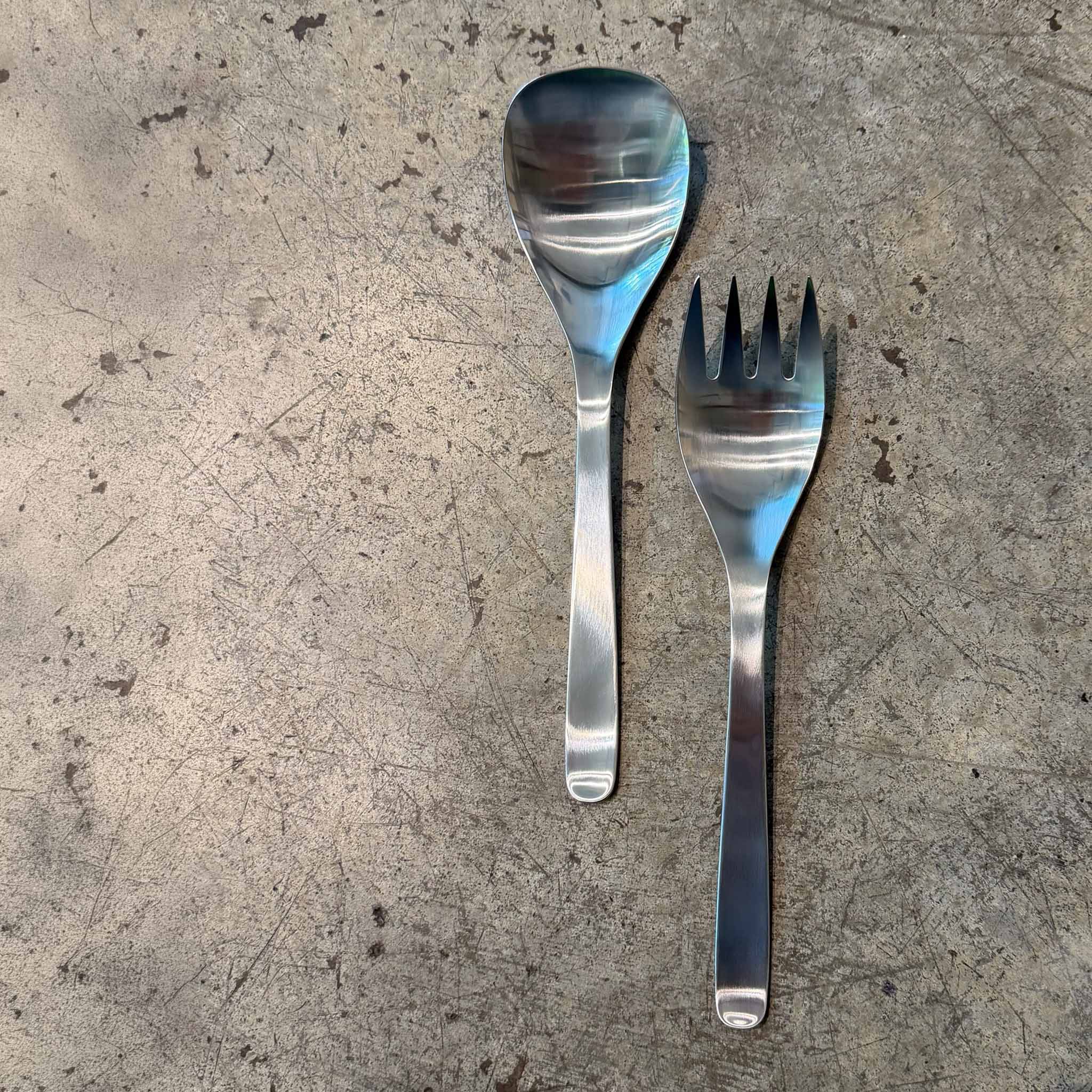 A stainless steel serving spoon and matching fork with a textured, brushed finish, laid flat on a gray concrete surface.