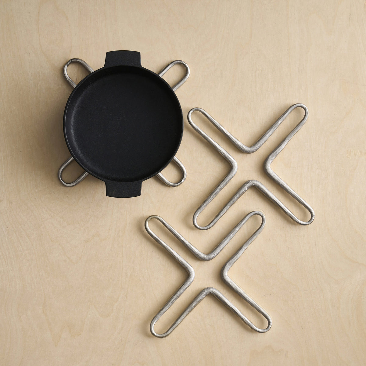 A black round cast-iron skillet with four loop handles sits on a light wood surface next to three matching silver metal trivet-style coasters.