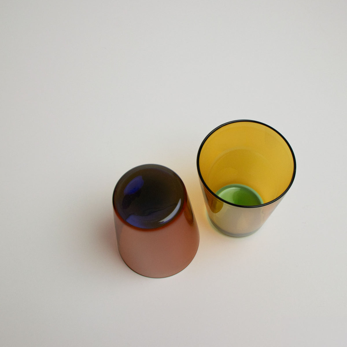 Two two toned stackable glass tumblers, one amber with a blue interior and one yellow with a green interior, on a white background.
