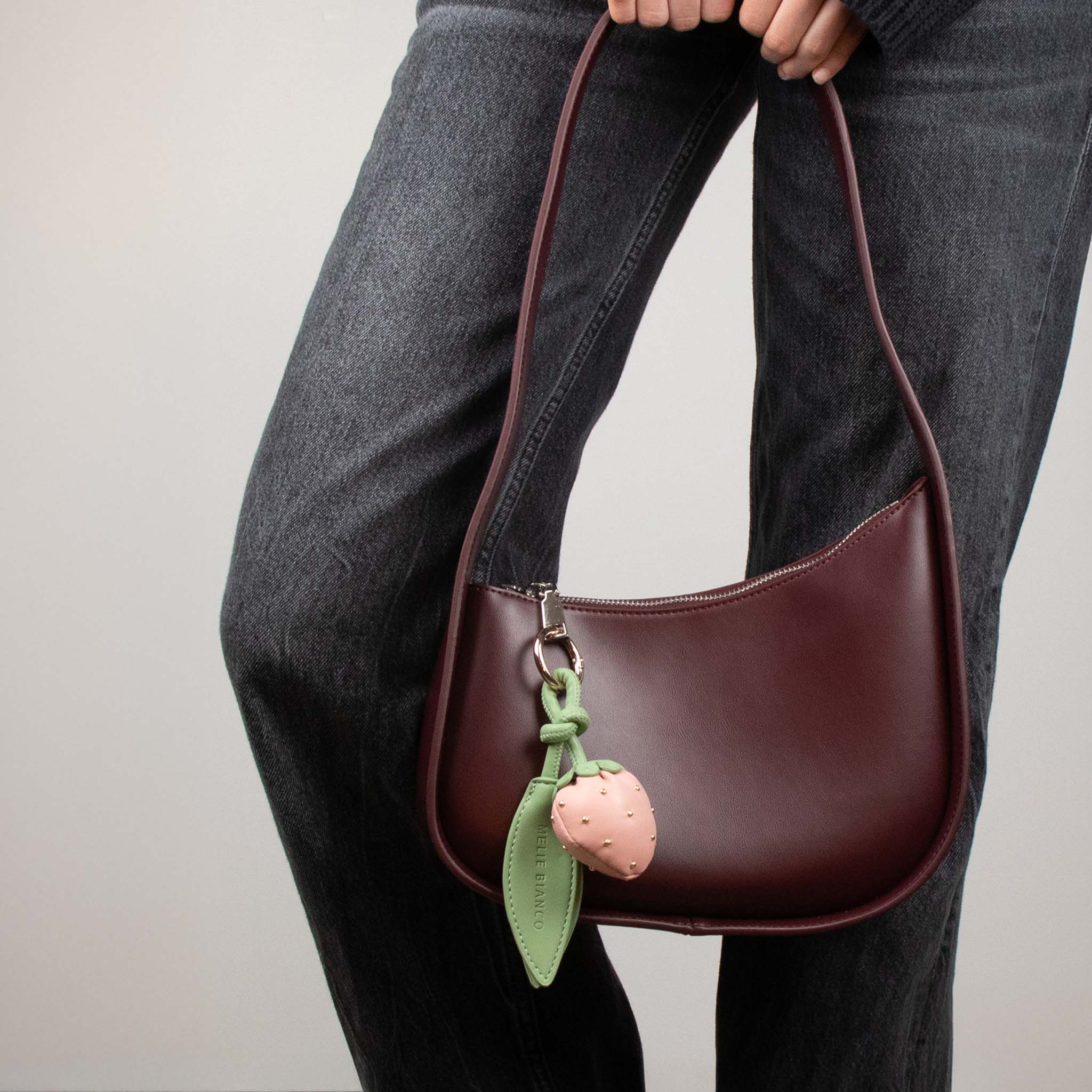 Image of model holding Melie Bianco's Shoulder Bag Willow in color plum adorned w/ a pink strawberry bag charm