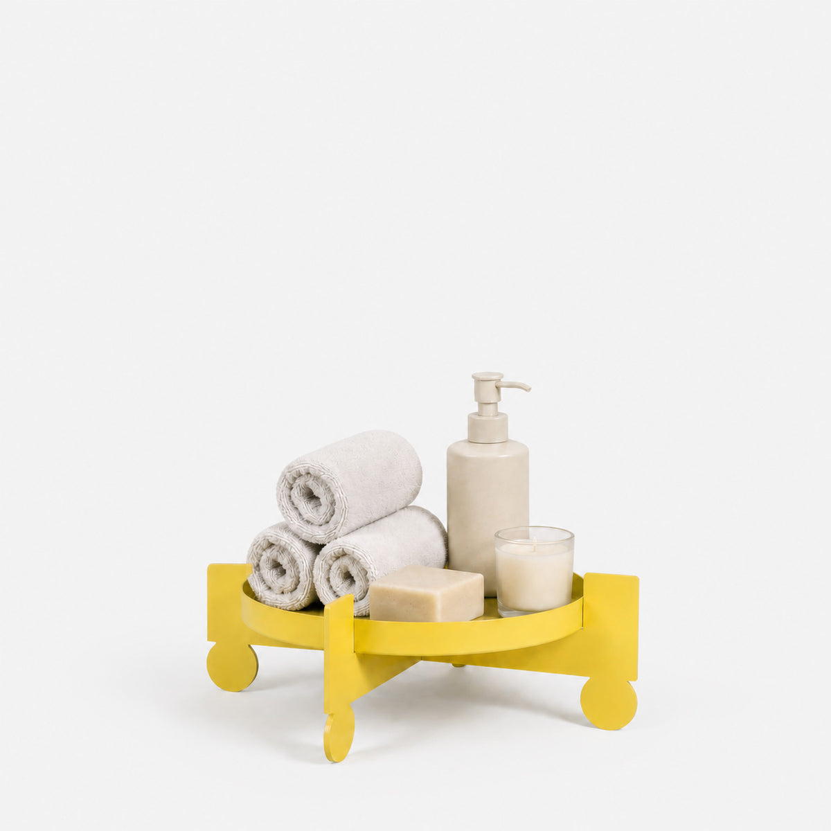 A bright yellow circular bathroom tray holding three rolled white towels, a beige pump bottle, a bar of soap, and a small candle, photographed on a clean white background.