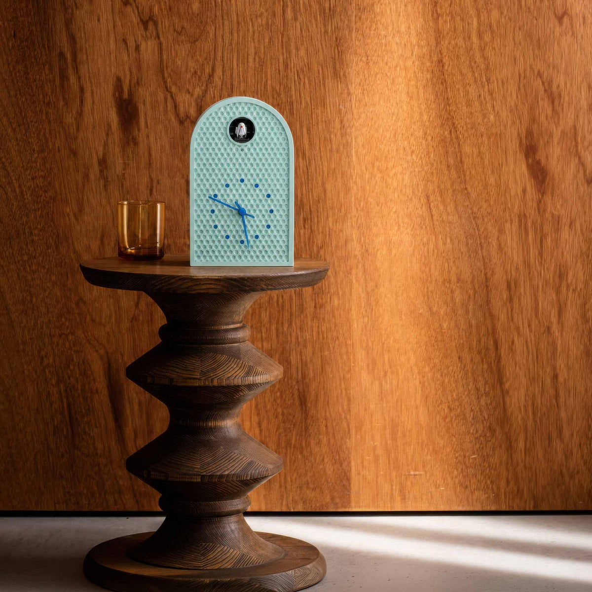 A mint blue arched cuckoo clock with a honeycomb pattern face and blue hands sits on a round wooden pedestal table against a warm wood-paneled wall. A small amber glass is also on the table.