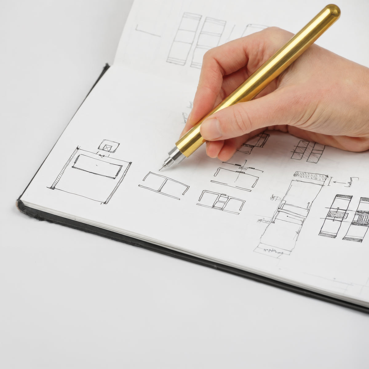 A close-up of a hand holding a metallic gold pen, sketching furniture plans in a white notebook.