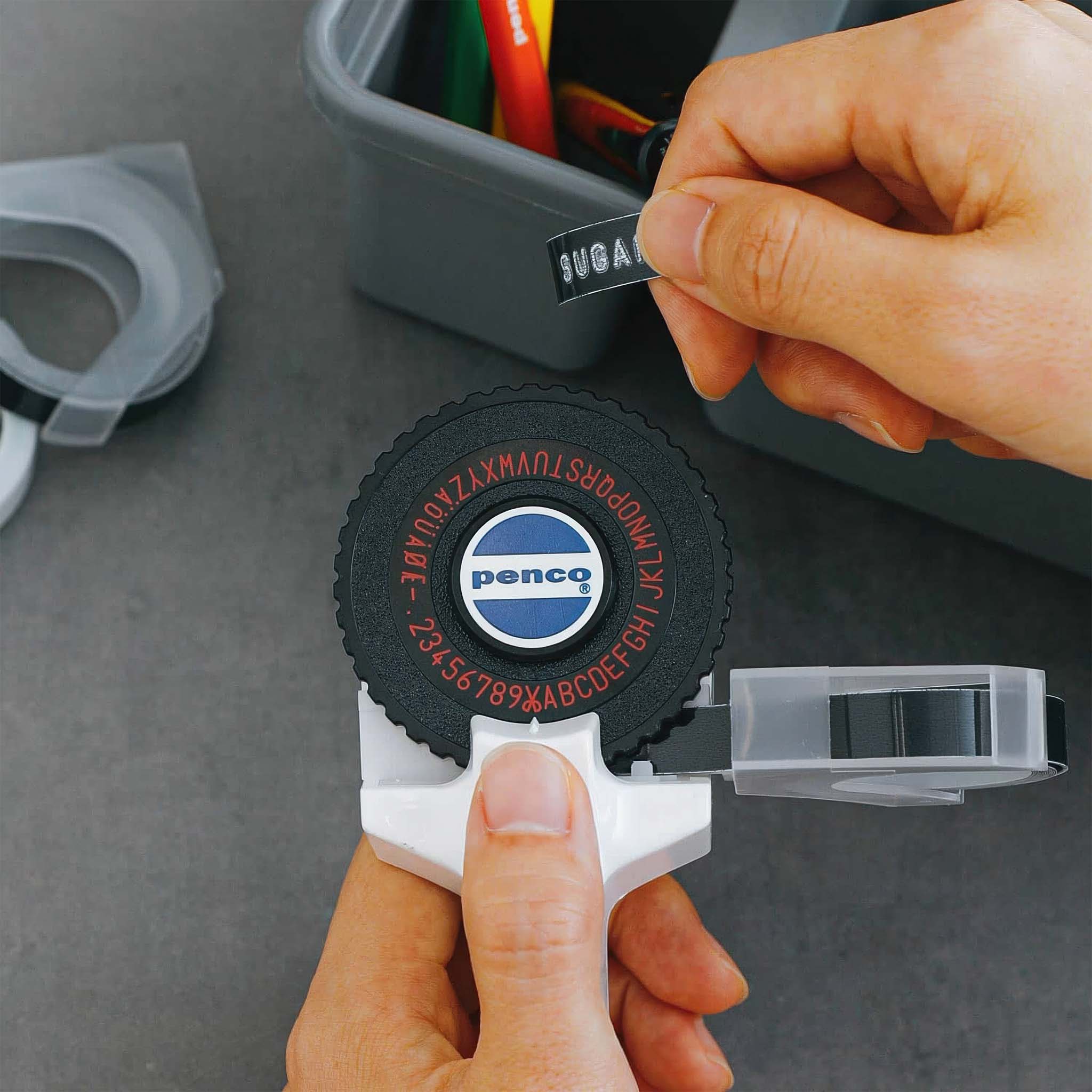 Person using a Penco label maker with a black label on a gray surface