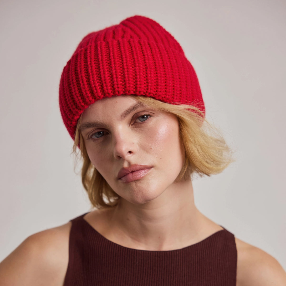 A close-up of a woman wearing a bright red ribbed knit beanie hat, paired with a dark brown ribbed tank top, against a neutral light gray background.