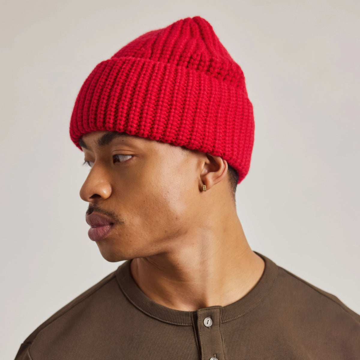 A man in profile wearing a bright red ribbed knit beanie and a brown henley shirt, photographed against a plain light gray studio background.