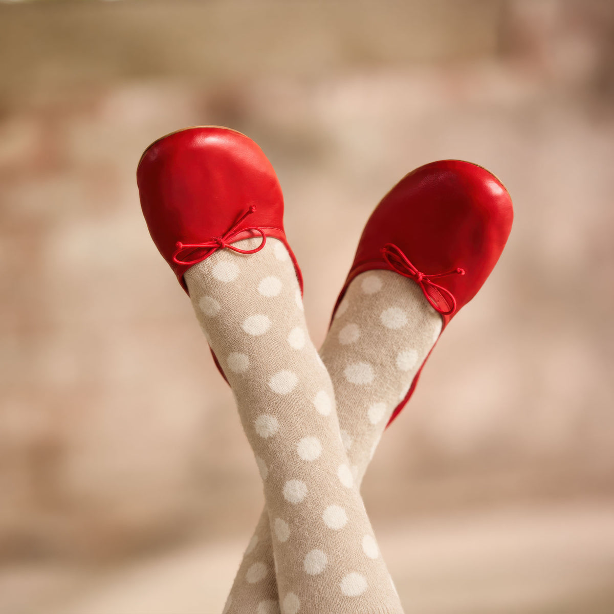 A close-up of crossed legs wearing lisa b's mushroom dot crew and shiny red ballet flats with red ribbon bows, against a softly blurred warm-toned background.