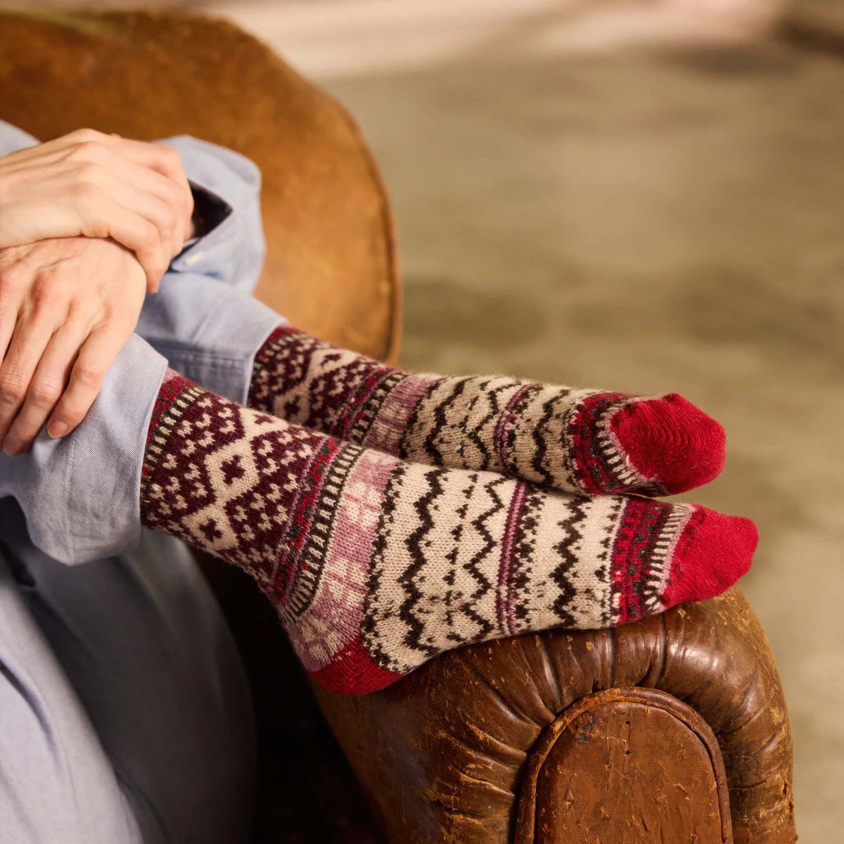 A person sitting in a brown leather armchair wearing a pair of lisa b's Red Fair Isle knit socks with red toes and heels, light blue pants, and crossed arms.