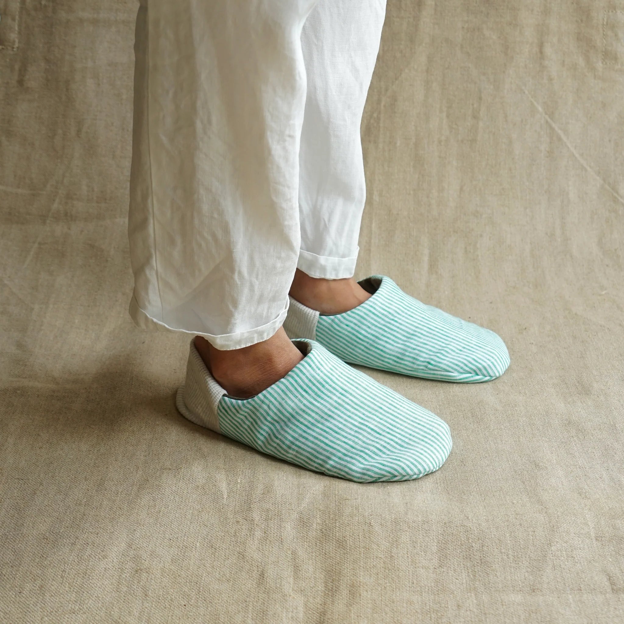 Person wearing a pair of mint green and white striped slippers with light grey heels on a beige fabric background