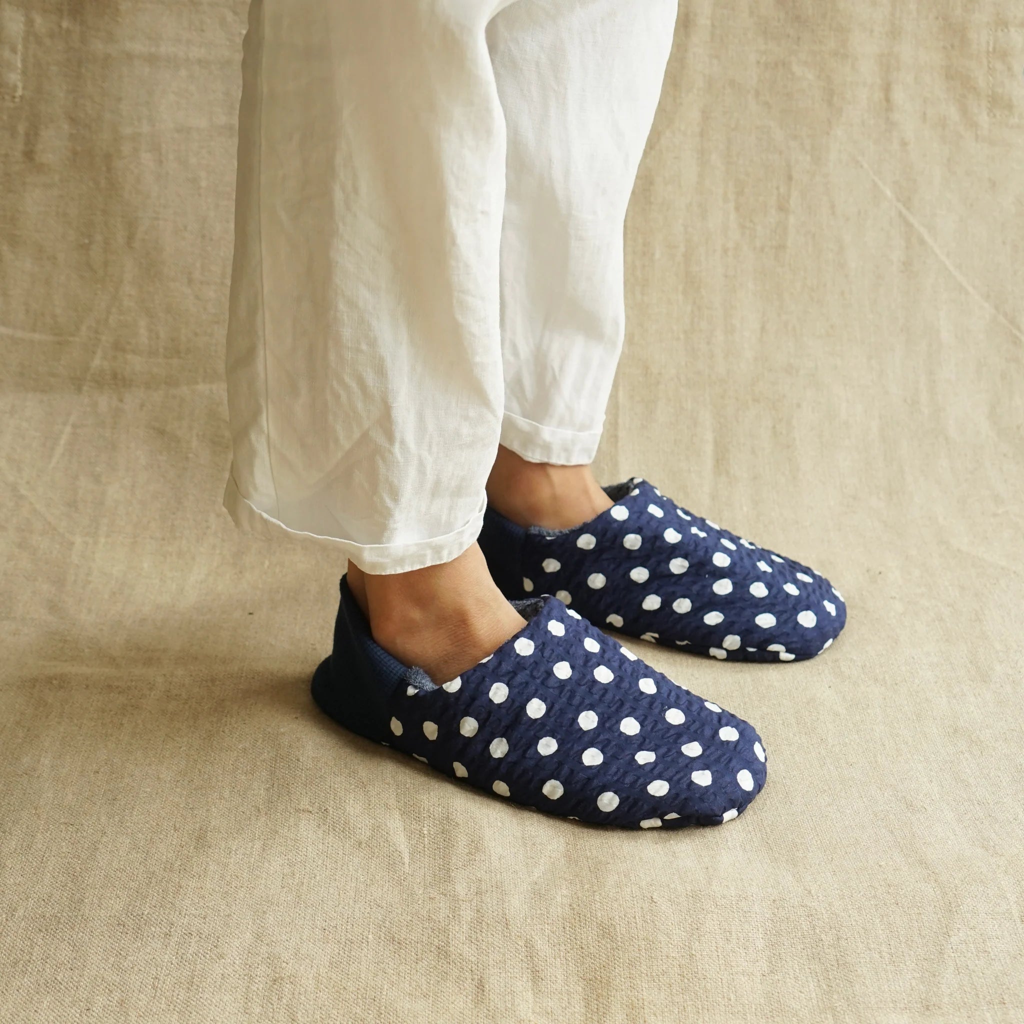 Navy blue slippers with white polka dots worn by a person in white pants on a beige background