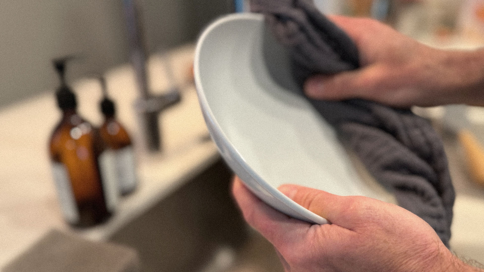 Hands drying a ceramic plate with a kitchen towel beside a sink.