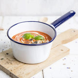 White enamel saucepan with blue rim containing tomato soup on a wooden board.