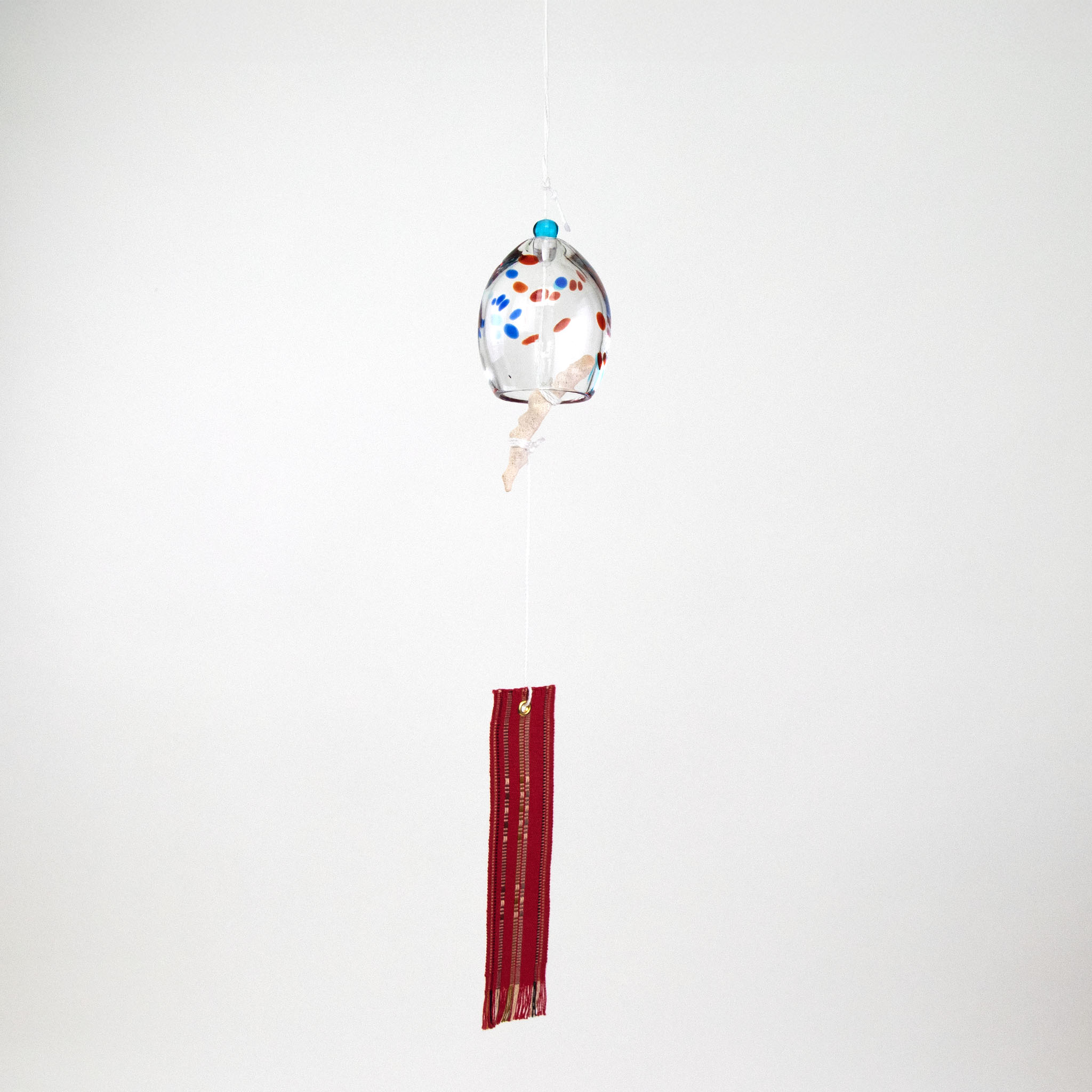 Colored speckled glass Okinawan wind chime with red handwoven tab by Miyahira Studio, suspended on a white background.
