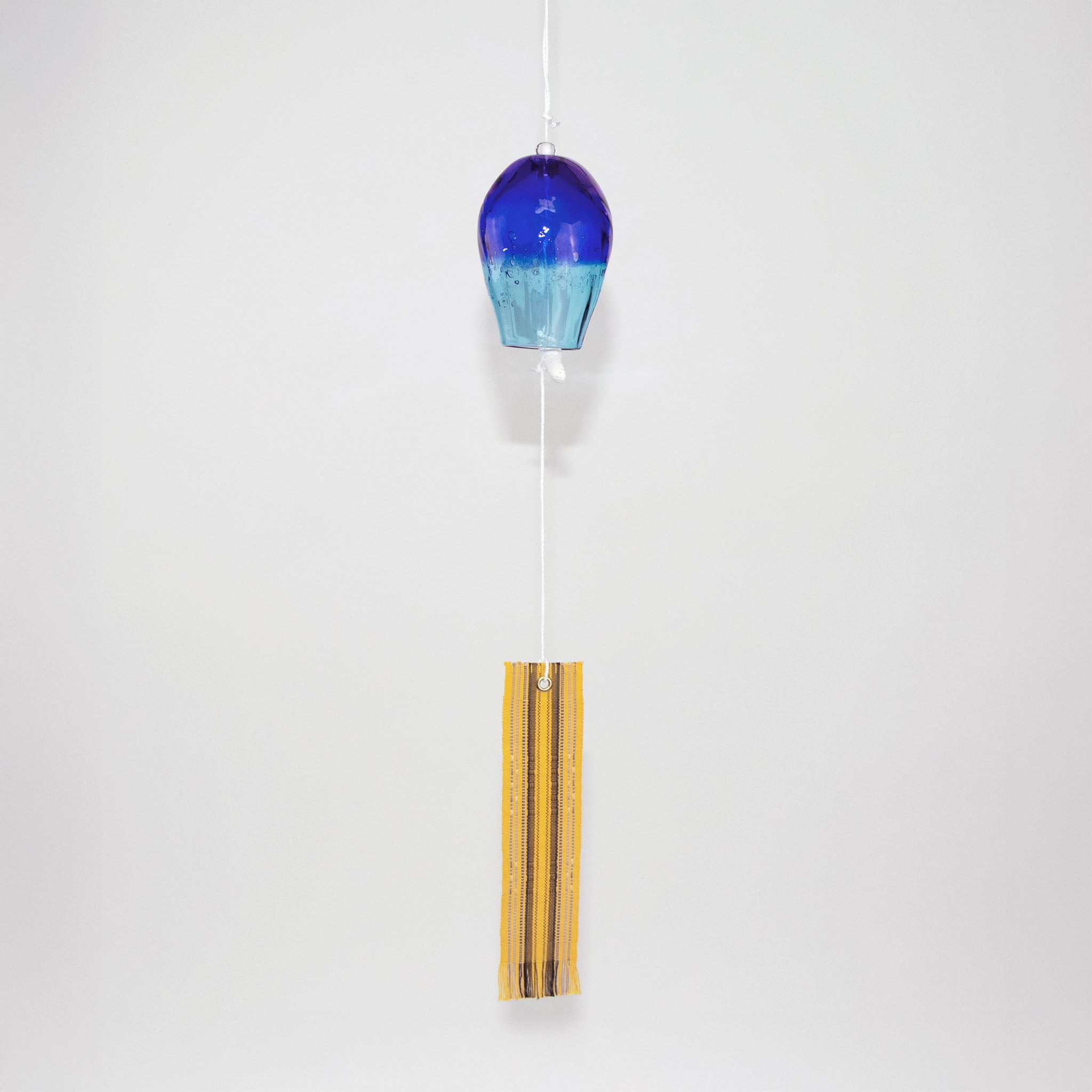 Blue glass Okinawan wind chime with yellow handwoven tab by Miyahira Studio, suspended on a white background.