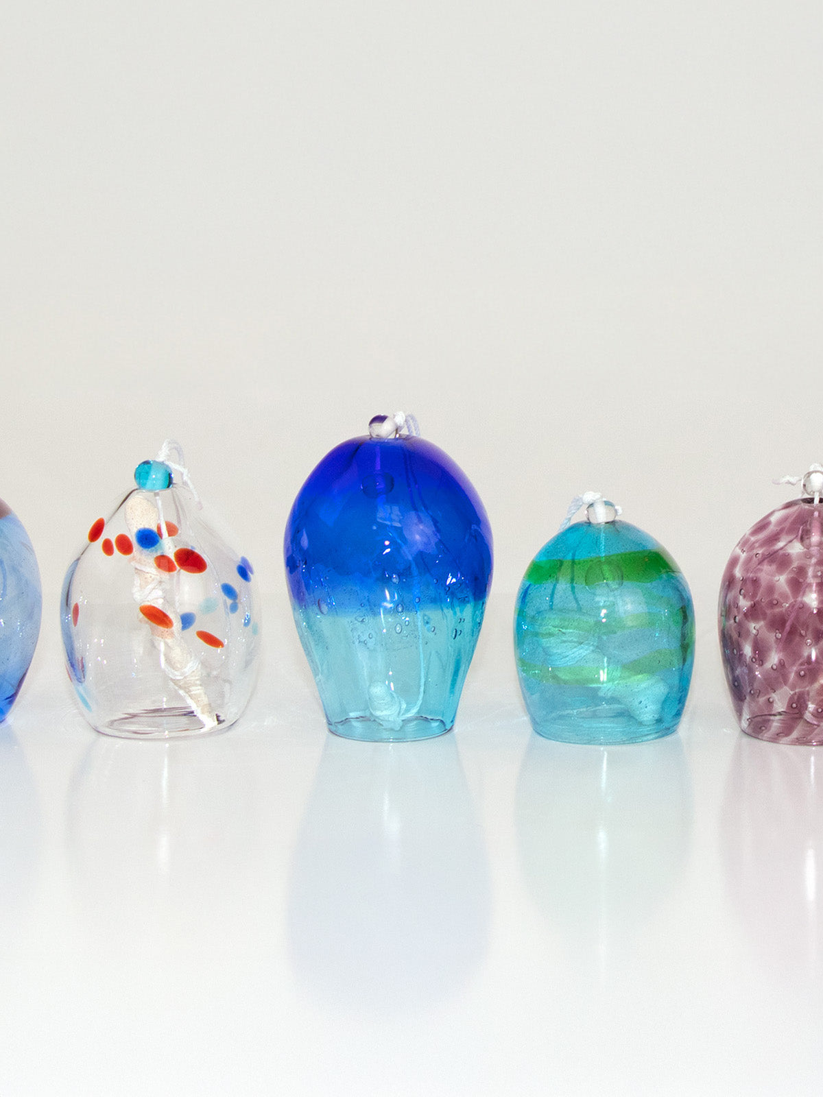 Eight handcrafted glass wind chimes from the Umikaze collection, arranged in a row and glowing in translucent shades of blue, aqua, green, and violet with wave-like and dotted patterns inspired by the sea breeze of Okinawa.