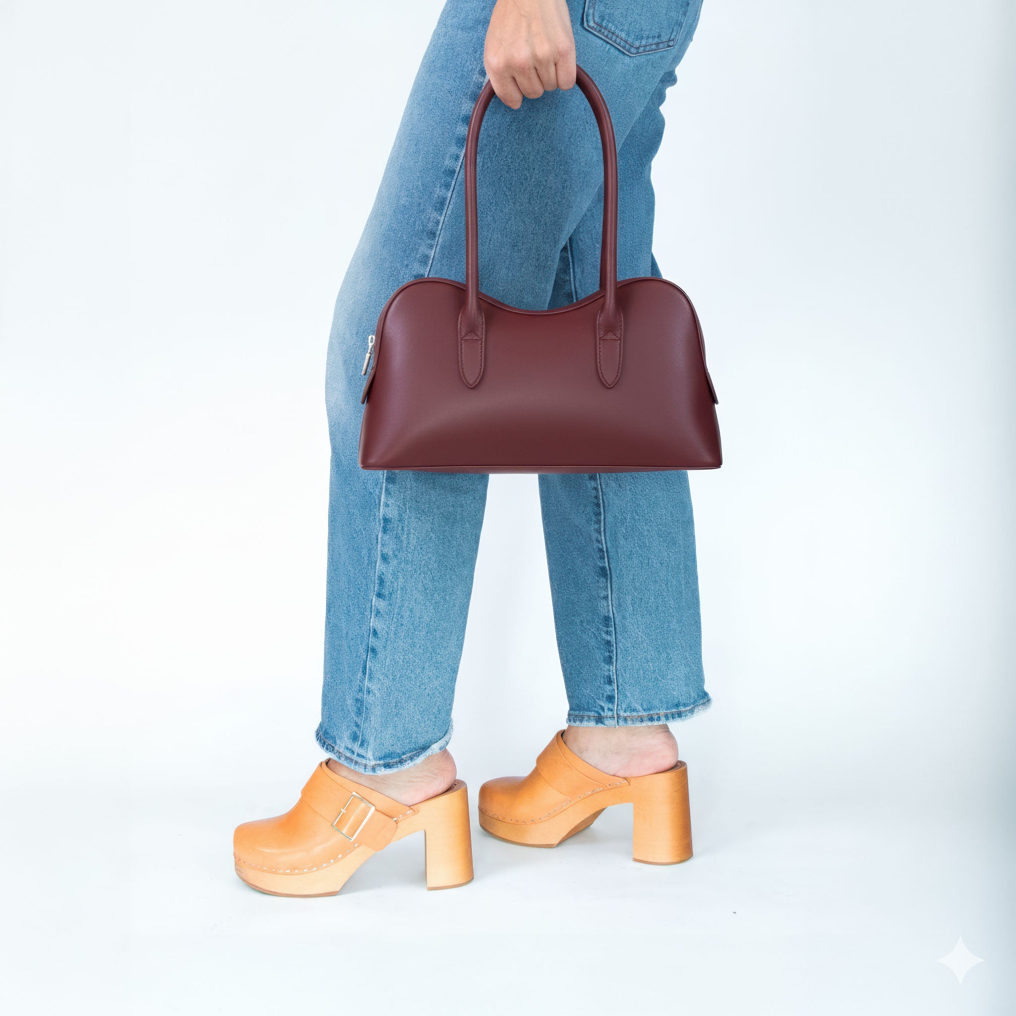 Person holding a maroon handbag with blue jeans and saddle colored shoes on a white background