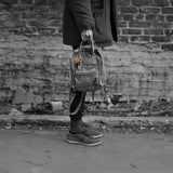 Person holding a backpack with CW&T's orange titanium key wrangler hanging off the handle against a brick wall.