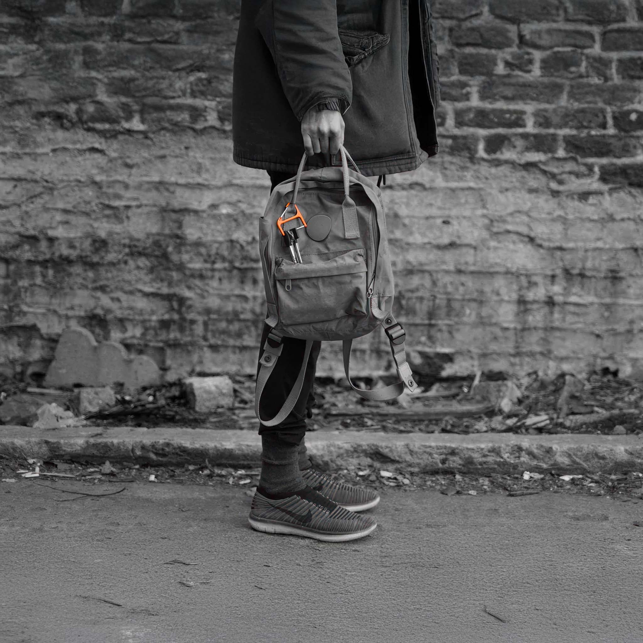 Person holding a backpack with CW&T's orange titanium key wrangler hanging off the handle against a brick wall.