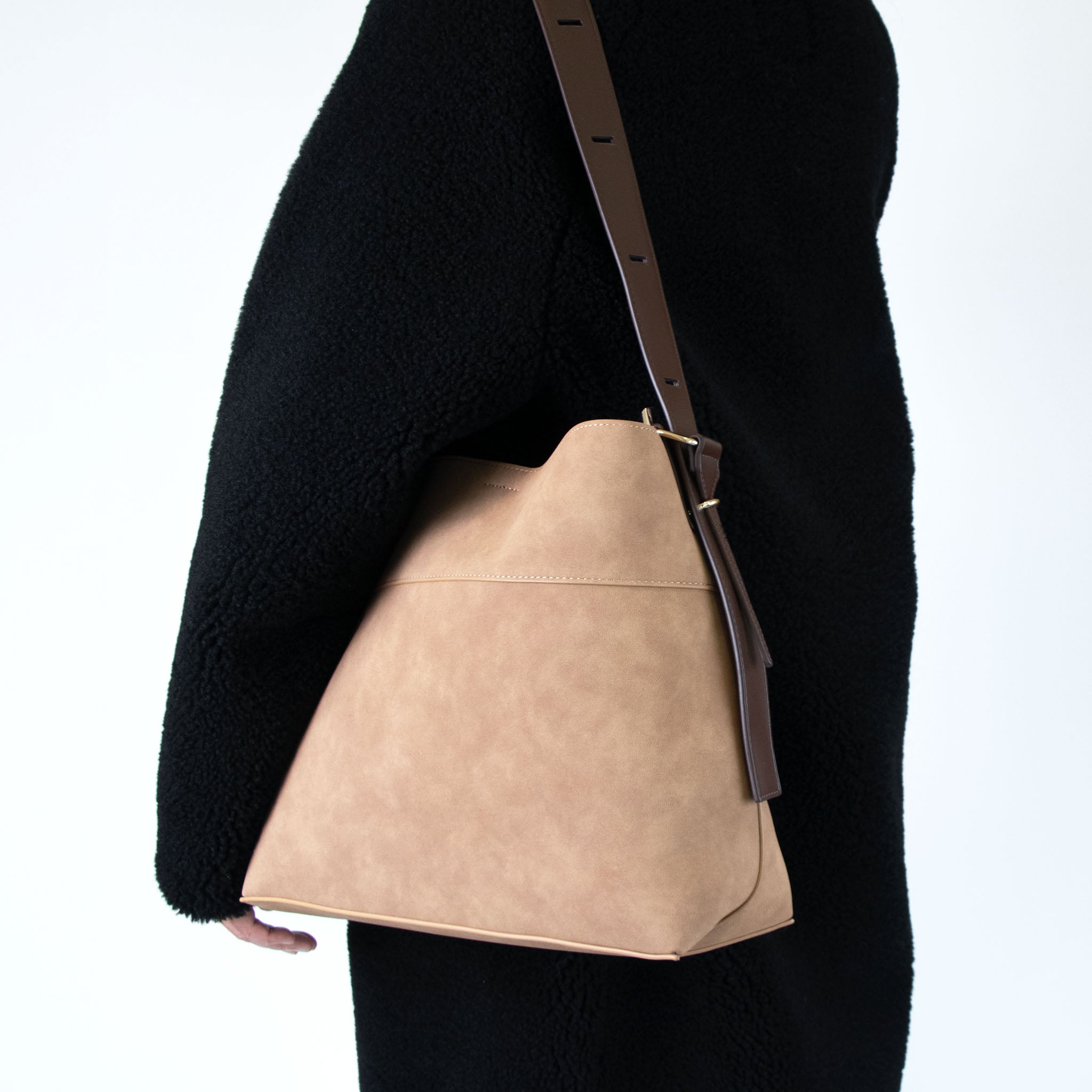 Mocha colored handbag with brown strap worn by a person in a black coat on a white background