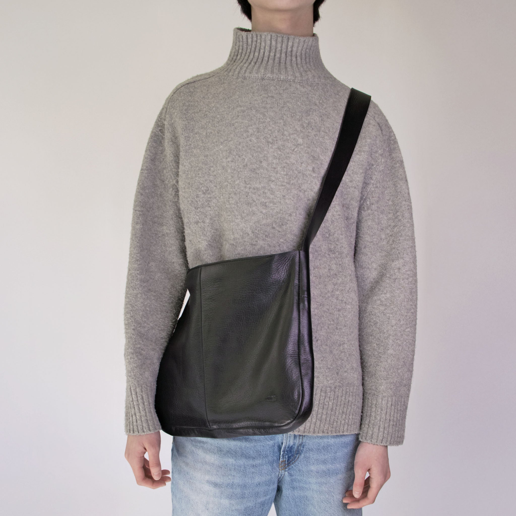 Person wearing a gray sweater and NImes black messenger bag on a plain background