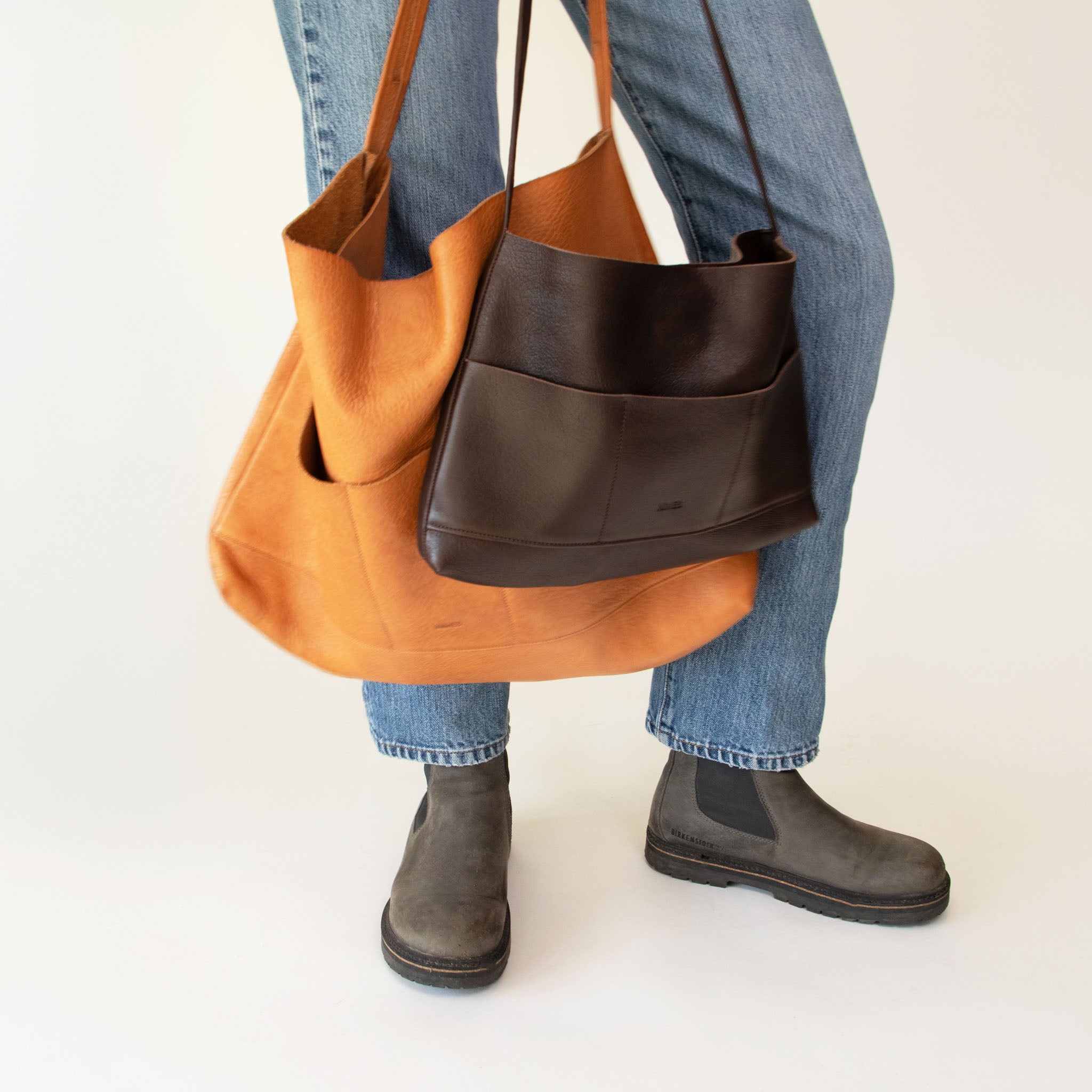 Person holding Nimes bonarda totes in both sizes while wearing blue jeans, and gray boots on a white background