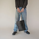 Person holding NImes black woven tote wearing blue jeans and black shoes on a plain background