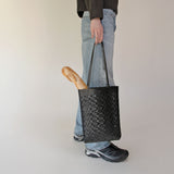 Person holding NImes black woven tote carrying a baguette and wearing blue jeans and black shoes on a plain background