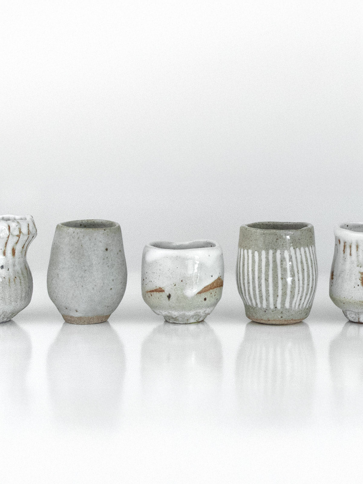 A row of ten one-of-a-kind handcrafted ceramic sake cups by The Lazy Potters, each with unique forms, glazes, and textures in soft white, grey, and earthy clay tones, displayed on a reflective surface.