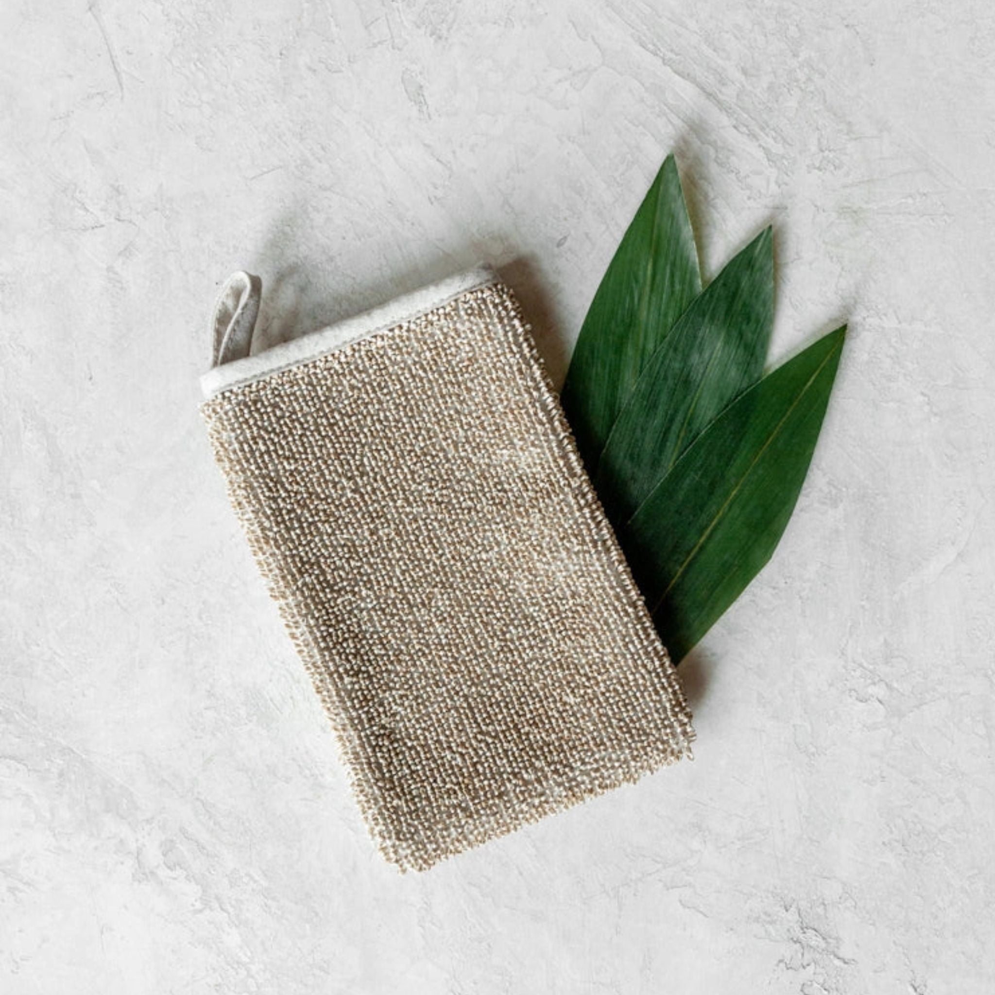 Textured Sasawashi body scrub mitt on a marble surface beautifully laid out on top of 3x green leaves