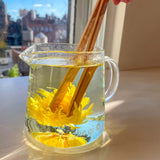Clear glass server with yellow flower tea and bamboo tongs on a reflective surface, blurred cityscape in the background.