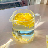 Clear glass server with yellow flower tea on a reflective surface, blurred cityscape in the background.