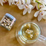 Tea cup with jasmine tea, jar labeled 'The Qi Single Origin Jasmine', and white flowers on a tan background