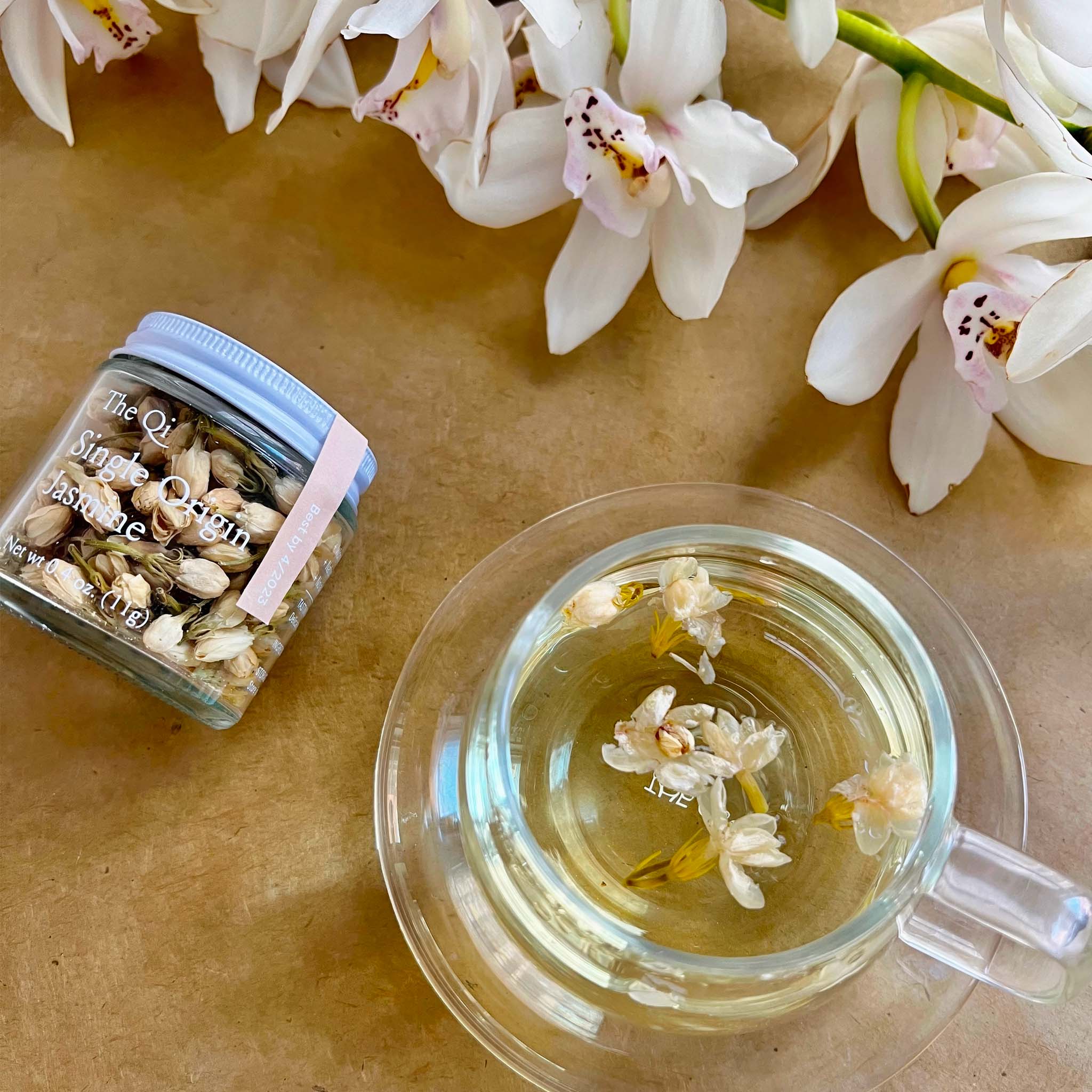 Tea cup with jasmine tea, jar labeled 'The Qi Single Origin Jasmine', and white flowers on a tan background